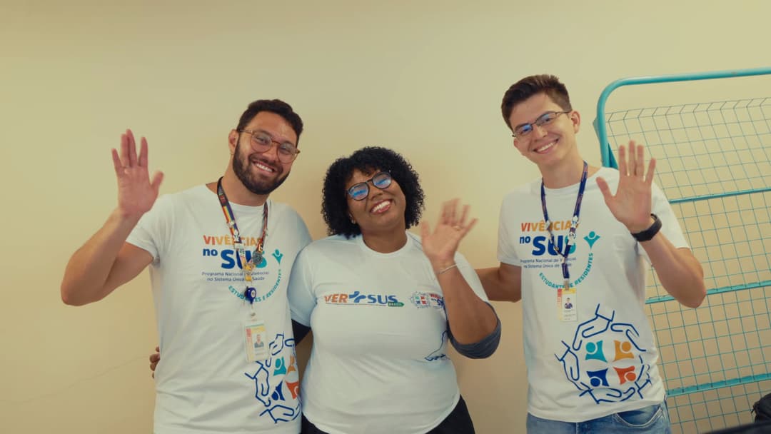 Três participantes com camisetas do Vivências no SUS acenam e sorriem para a câmera.
