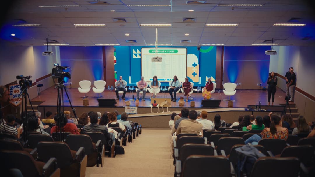Auditório visto do fundo, com duas câmeras em tripé no corredor lateral e a mesa no palco.