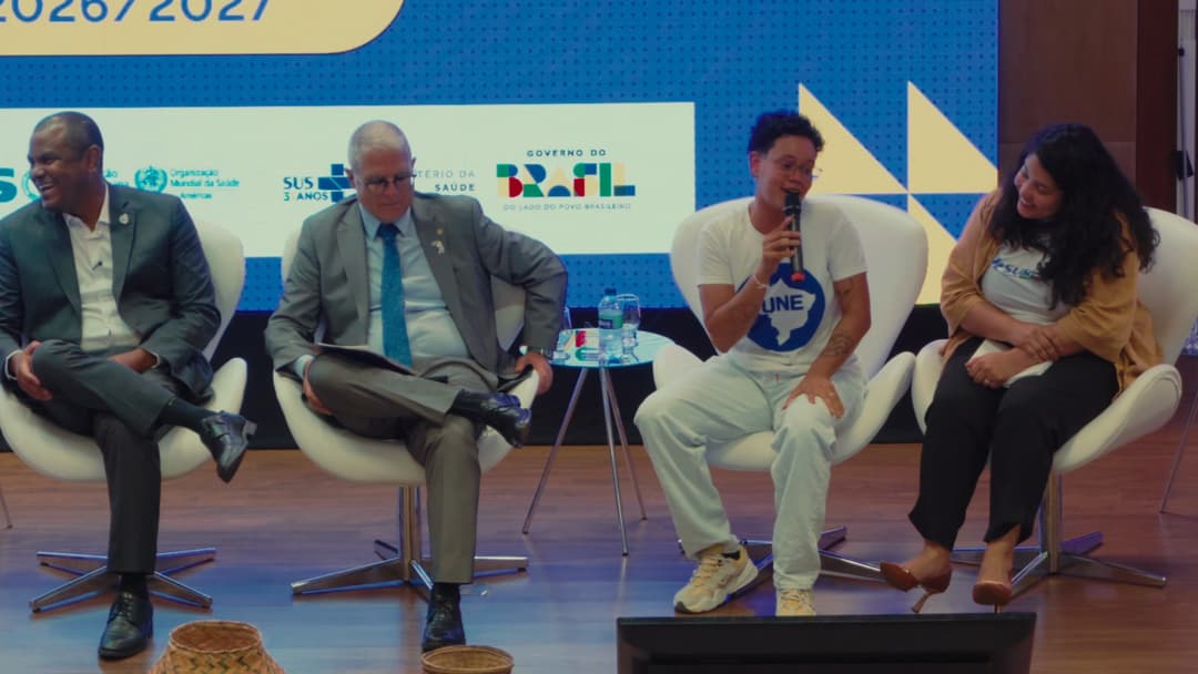 Quatro integrantes da mesa sentados no palco; um jovem com camiseta da UNE fala ao microfone e os vizinhos sorriem.