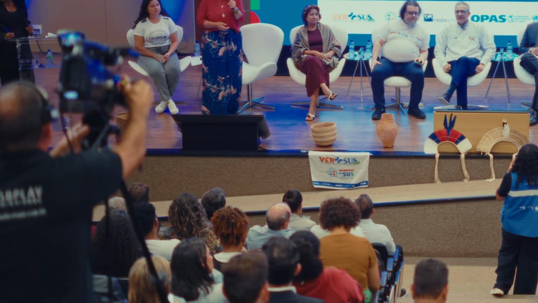 Cinegrafista de costas, com câmera no ombro, filma a mesa no palco do auditório.