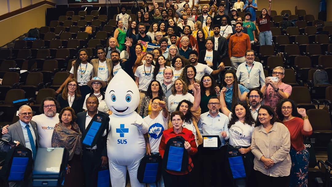 Foto coletiva no auditório com o mascote Zé Gotinha, integrantes da mesa e o público de pé.