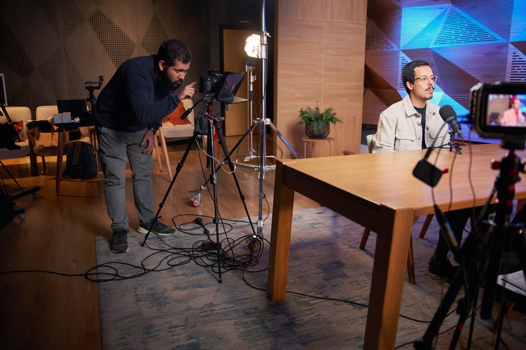 Eduardo Braz, da LENZ, ajusta câmera e teleprompter enquanto Guilherme Alves aguarda sentado à mesa