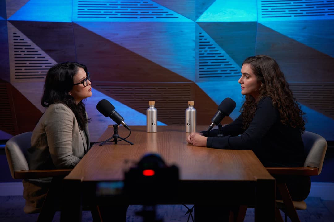Bianca Orrico e Isabella Ferro conversam à mesa com microfones, com a câmera gravando em primeiro plano