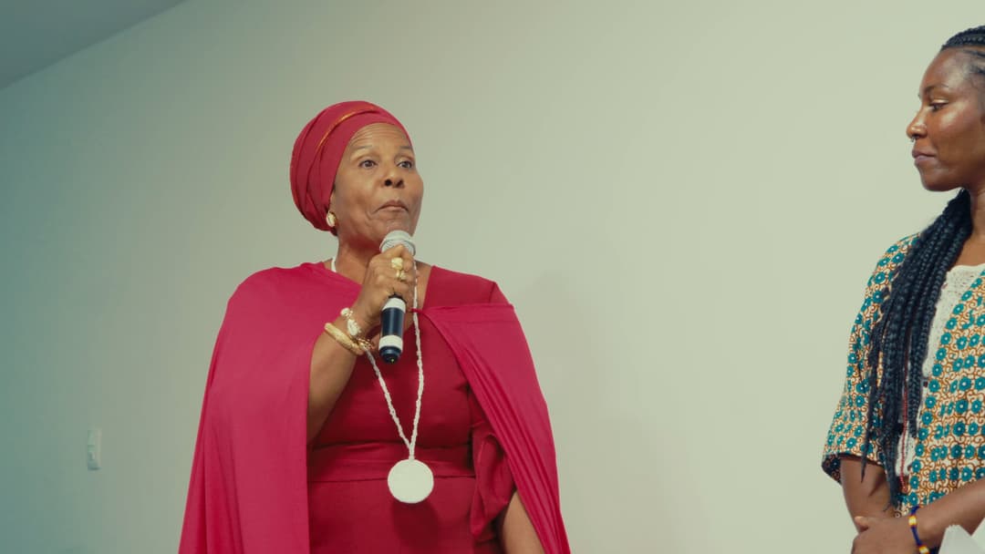 Mulher de turbante e capa vermelha segura o microfone, observada por outra participante.