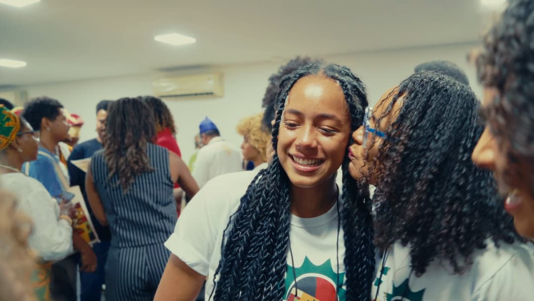 Duas participantes com camisetas do programa se cumprimentam; uma sorri enquanto recebe um beijo no rosto.