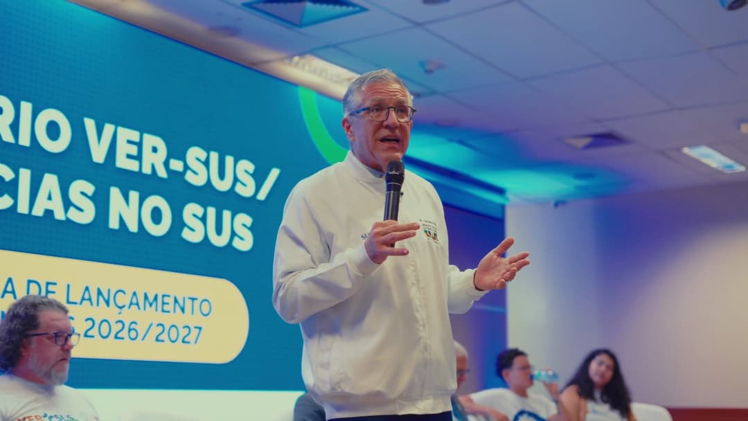 Frame do vídeo C7HkBSB2LtY O Ministro da Saúde fala ao microfone, de pé, diante do painel do Seminário VER-SUS.