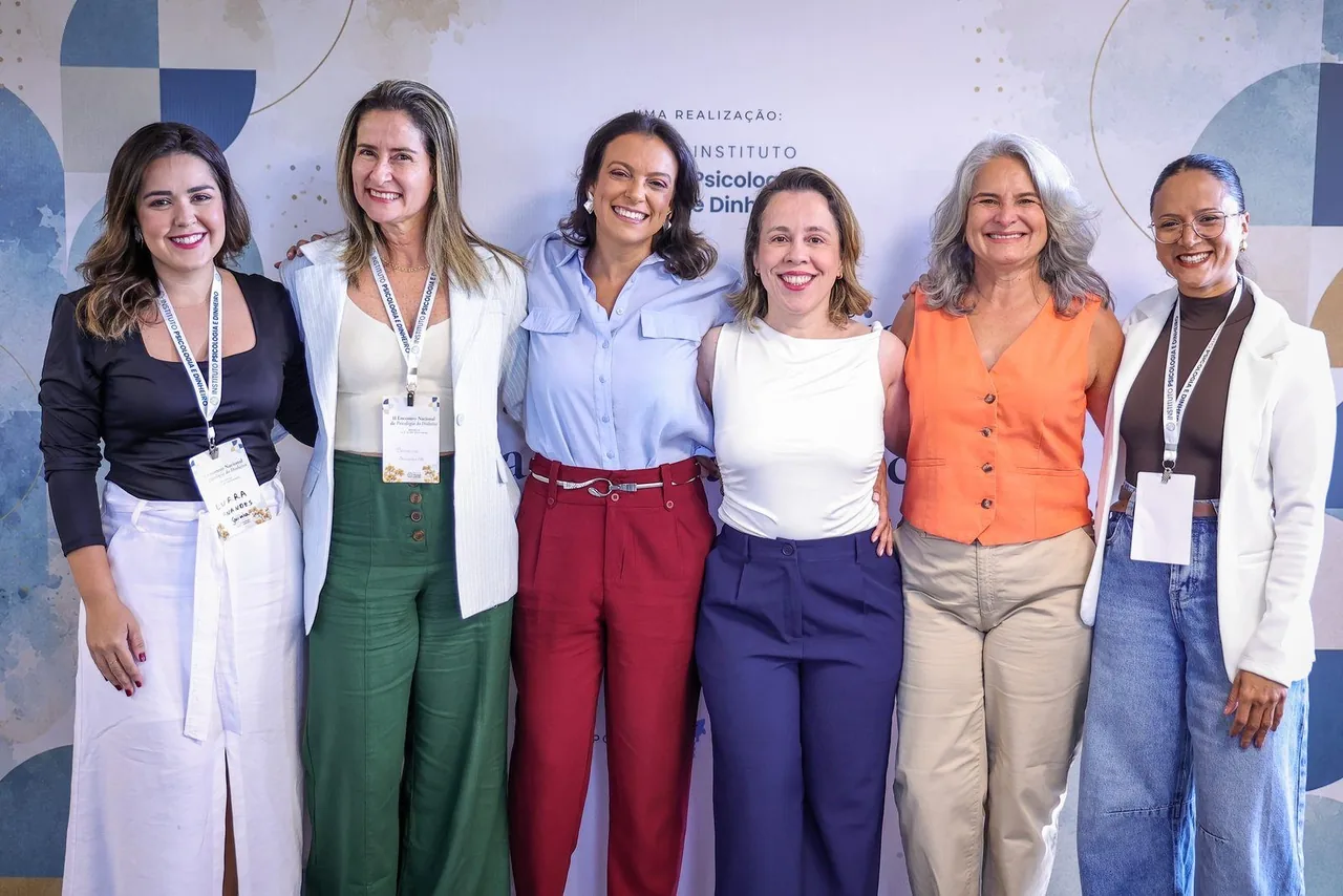 Seis participantes abraçadas lado a lado diante do painel do II Encontro de Psicologia do Dinheiro