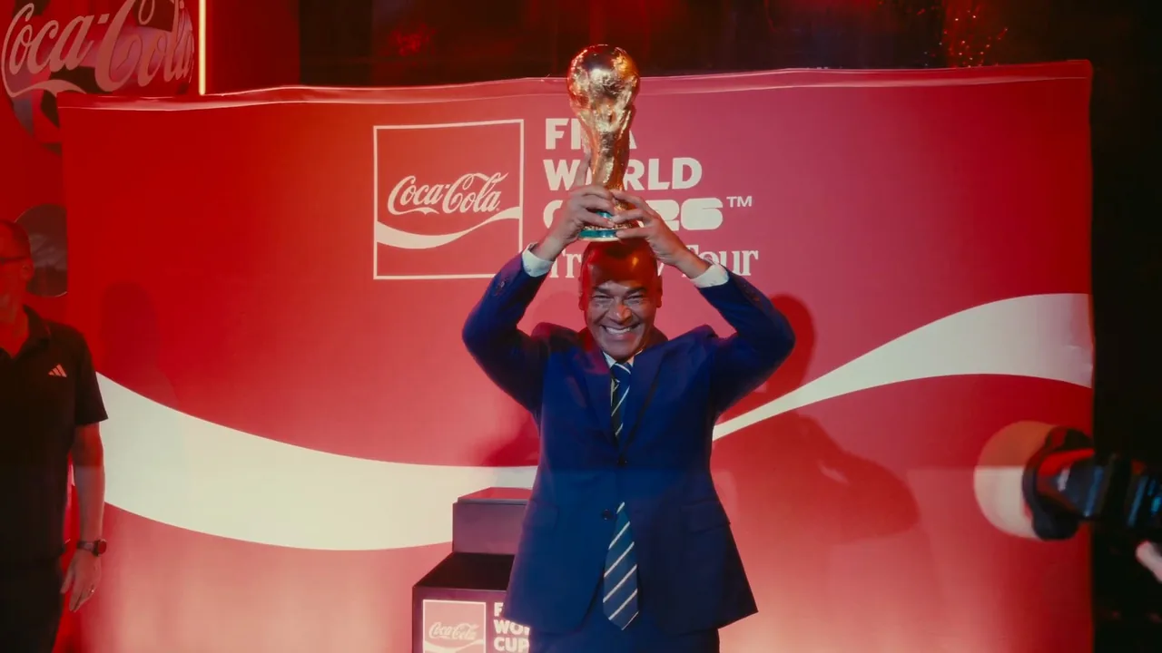 Homem de terno levanta a taça da Copa do Mundo diante de um painel vermelho do Trophy Tour