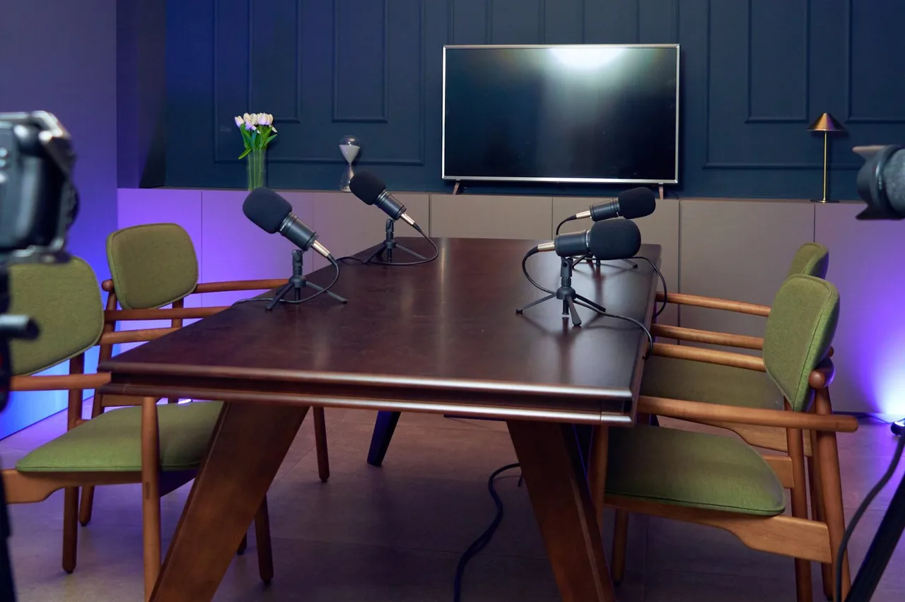 Mesa de madeira do estúdio da LENZ com quatro microfones de mesa em pedestais, cadeiras estofadas ao redor e televisão na parede ao fundo